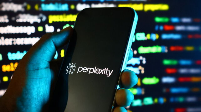Smartphone displaying perplexity logo held in hand with computer code background lighting.