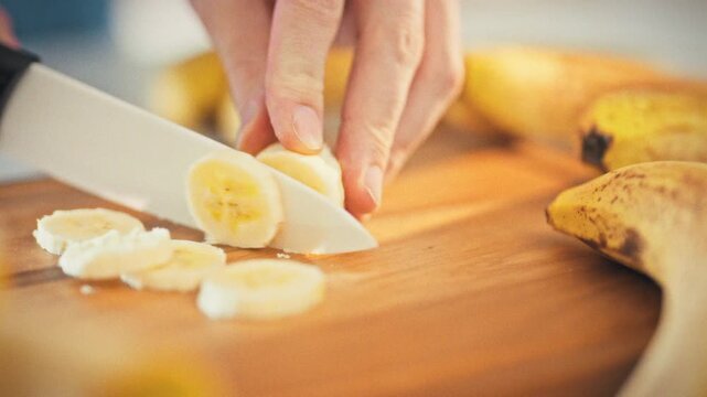 A man slices a ripe banana with a ceramic knife on a cutting board, monitoring his diet. The scene was filmed in a home kitchen on a bright sunny day. The concept is diet food