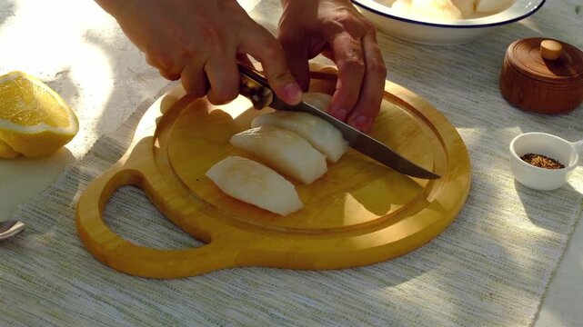 A chef cuts cod or other white fish fillets into strips on a board against a white background outdoors. Step-by-step video recipes. Ingredients for cooking