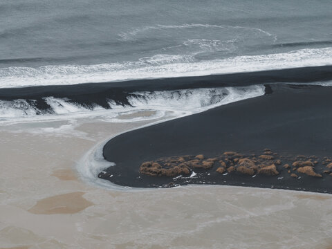 Aerial view of the black sand beach with white sea foam, crashing waves, and patches of dry grass in Reykjavik, Reykjavikurborg, Iceland.