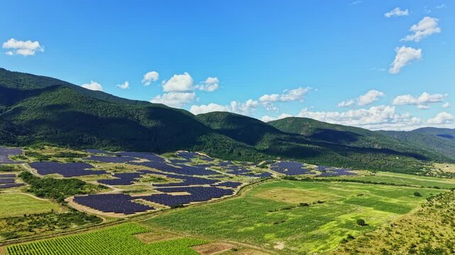 Solar panels are arranged in rows across wide fields in a rural area with green land and mountains in the background. The sky is blue with some clouds.