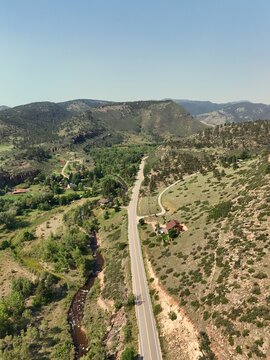 Aerial view of a winding road through a lush green valley with rocky hills, a flowing stream, and scattered trees near Hooter Lookout in Lyons, Colorado, United States.