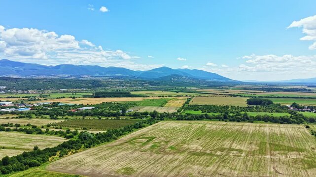 Fields stretch out across the landscape with mountains rising in the background under a bright sky. Farmers may be working the land or preparing for harvest.