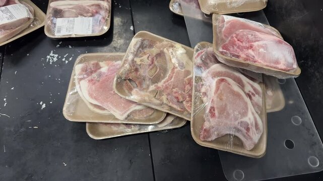 Packaged pork chops lie in a disordered stack on a black supermarket display shelf, wrapped in clear plastic on cardboard trays. The untidy arrangement suggests retail merchandising or hygiene concern