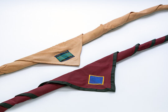 Two Thai scout neckerchief scarves in tan beige and dark maroon red colors featuring blue and tartan plaid fabric patches with rolled woggle knots arranged neatly on clean white studio background