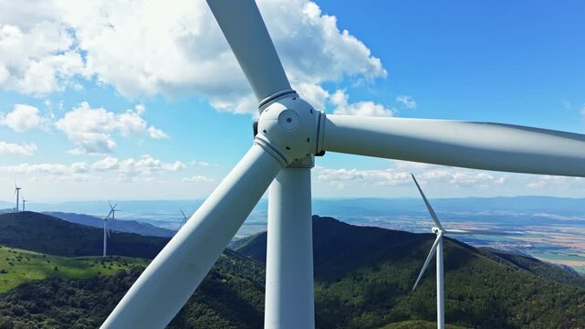Wind turbines are situated on a green hilltop under a blue sky with clouds. The scene shows a landscape where wind energy is produced. Turbines spin slowly, taking advantage of the wind.