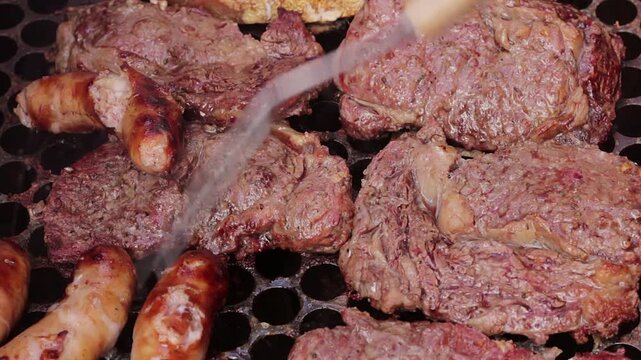 Beef steaks and sausages being turned with a large fork on a hot grill