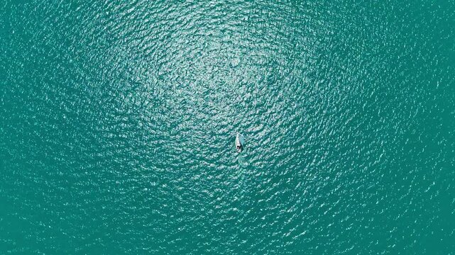 A small sailboat moves through clear blue waters. The sun shines brightly over the ocean, creating patterns on the surface. The scene captures a moment of sailing and leisure.