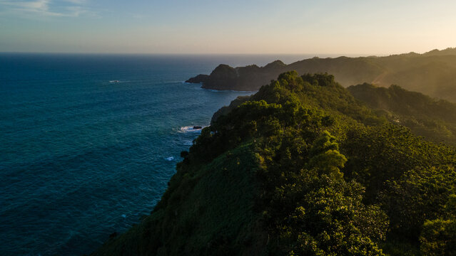 Aerial view of the lush green cliffs meeting the deep blue ocean at sunset in Kebumen, Central Java, Indonesia.