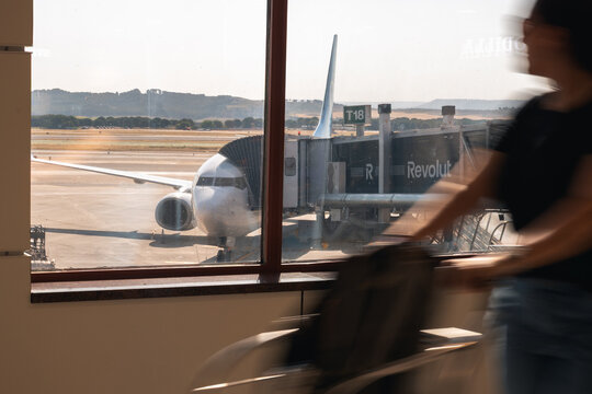 Passenger walking past airport terminal window with parked airplane and jet bridge at international airport in Madrid, Spain - June 8, 2026