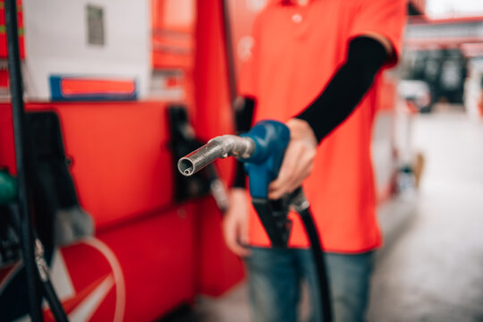close-up, selective-focus shot at a gas station. An attendant, wearing a bright orange polo shirt and a black arm sleeve, holds out a fuel pump nozzle with a blue handle