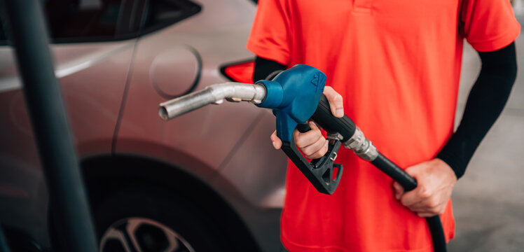 close-up, selective-focus shot at a gas station. An attendant, wearing a bright orange polo shirt and a black arm sleeve, holds out a fuel pump nozzle with a blue handle
