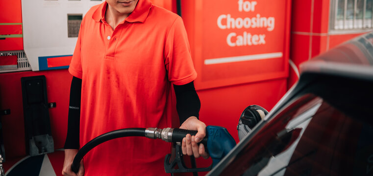a male gas station attendant in the process of refueling a vehicle.He is looking down attentively as he holds a blue-handled fuel nozzle inserted into the open fuel tank of a dark-colored car.
