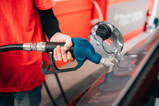 a close-up view of a person refueling a vehicle. The focus is on the individual's hand gripping a blue-handled fuel pump nozzle that is inserted into the open fuel tank of a dark car.