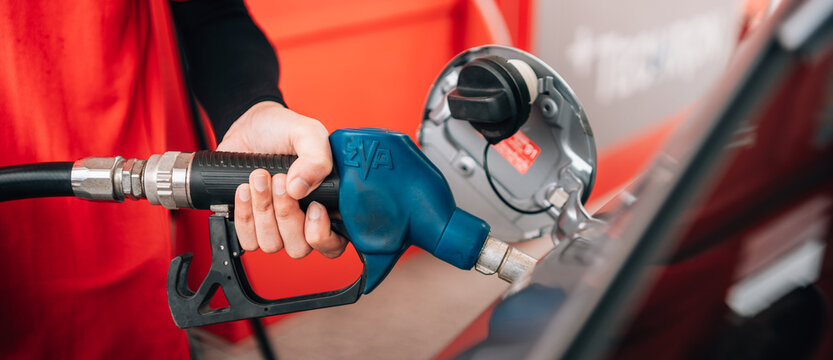 a close-up view of a person refueling a vehicle. The focus is on the individual's hand gripping a blue-handled fuel pump nozzle that is inserted into the open fuel tank of a dark car.