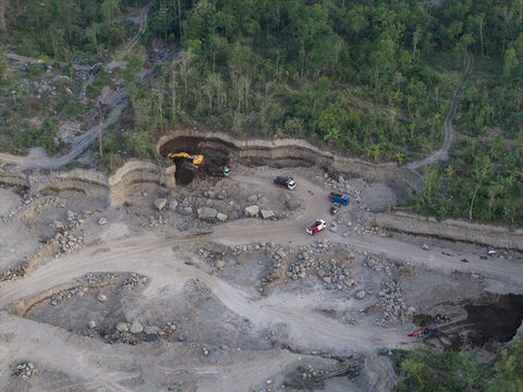 Aerial view of Sand and Stone Quarry with an excavator and trucks loading materials in a rugged landscape surrounded by lush forest in Manisrenggo, Central Java, Indonesia.