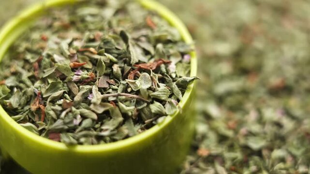 Organic dried oregano flakes falling in green plastic jar lid. Scattered herbs in the background. Culinary spice. Rotation