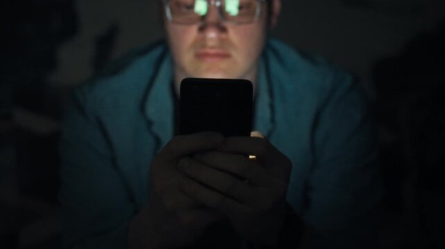 In a dark bedroom, a man glances at his smartphone's bright screen, habitually checking it late. This modern tech moment underscores digital addiction and its disruptive effect on sleep
