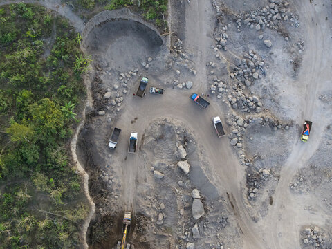 Aerial view of a sand and stone quarry with trucks and heavy machinery operating on dirt roads surrounded by lush vegetation in Manisrenggo, Central Java, Indonesia.
