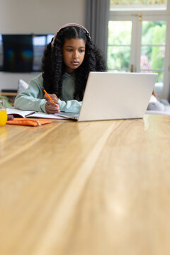 African American teen studying at home on table using laptop, taking notes, wearing pink headphones