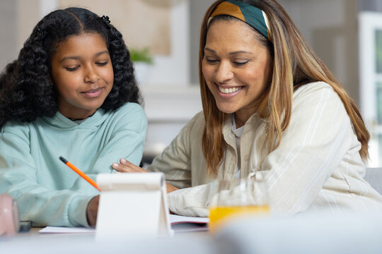 African American mom helping adolescent daughter at table, writing with orange pencil by flip stand