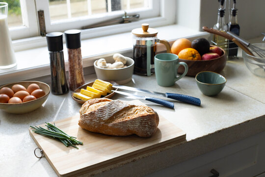 Crusty artisan loaf resting on wooden cutting board with chives on countertop, eggs, coffee brewing