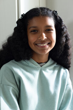 African American girl smiling near window at home, wearing mint hoodie, black clips, necklace