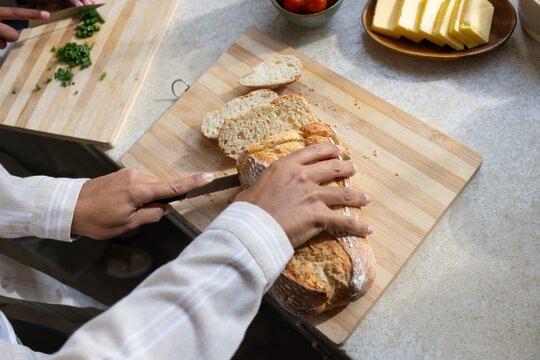 Diverse cooks slicing rustic loaf on counter while woman in light shirt, assistant chopping herbs