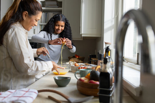 African American mother and teen daughter whisking with whisk, cracking eggs at kitchen counter
