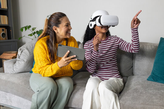 African American mother and daughter using VR sitting on grey sofa holding tablet