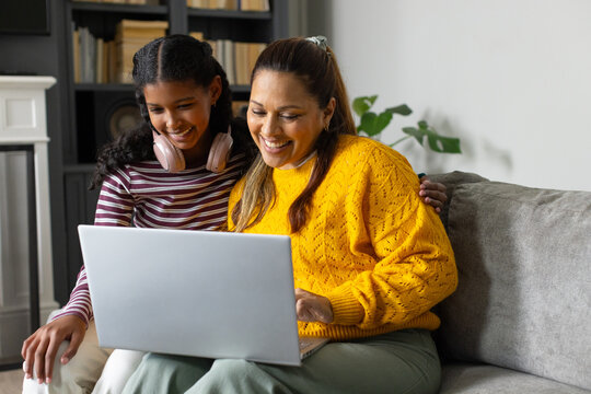 African mother in mustard sweater and daughter with pink headphones sharing laptop on gray sofa