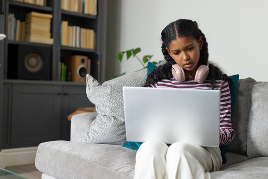 Teenage African female studying on sofa in striped top, using laptop with pink headphones