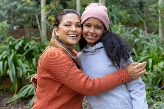 African American mother and teenage daughter hugging and smiling in garden wearing rust sweater