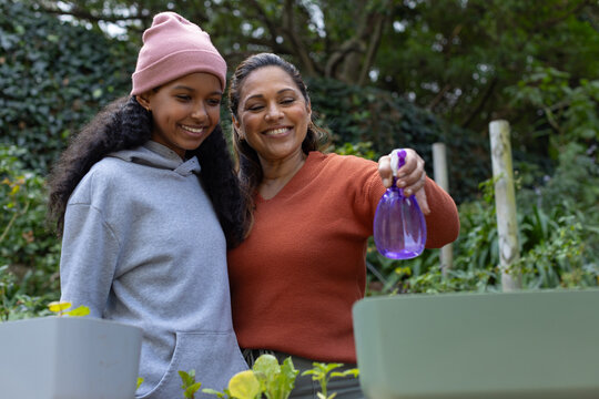 African American mother holding purple sprayer, teenage daughter in beanie misting garden planters