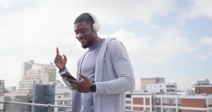 African man on rooftop glancing phone, tapping, controlling music with headphones, nodding, smiling