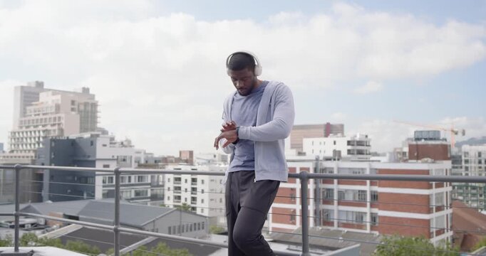 Mid-adult man checking watch, leaning on rooftop, tracking workout, moving to music on headphones