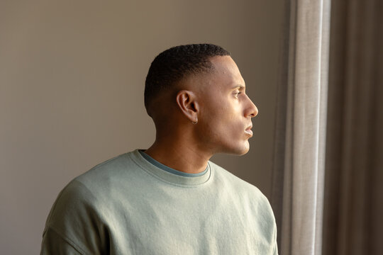 Adult African American man posing by window with curtains in light-green crewneck and hoop earring