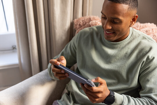 Man holding tablet in protective case sitting in armchair near window wearing sage-green sweatshirt