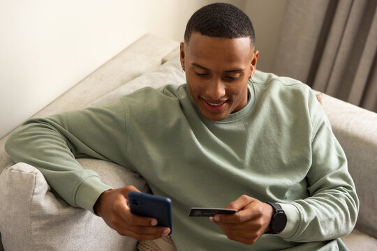 Adult African American man sitting on sofa at home wearing dark watch holding blue smartphone, card