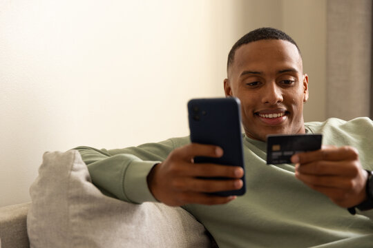 African American man reclining on sofa in living room, holding smartphone and black payment card