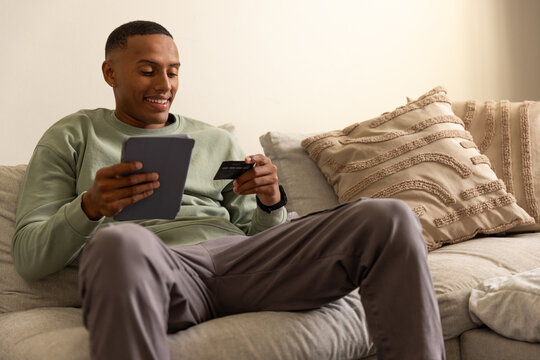 African American man sitting in living room on sofa holding tablet and credit card, smiling