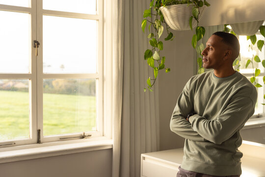 Adult African man standing at home near multi-pane window with latch, gazing out, copy space