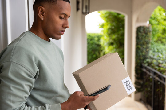 African American man in his twenties checking smartphone while holding barcode box on covered porch