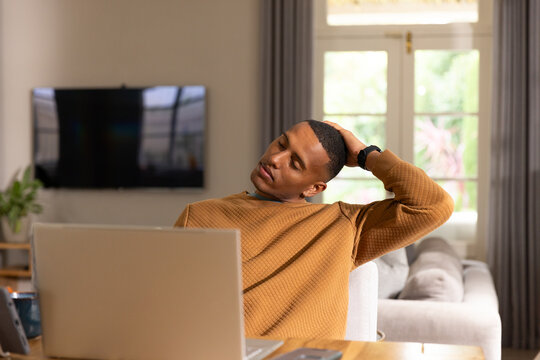 African man stretching at wooden table in home workspace wearing mustard sweater, laptop, mug