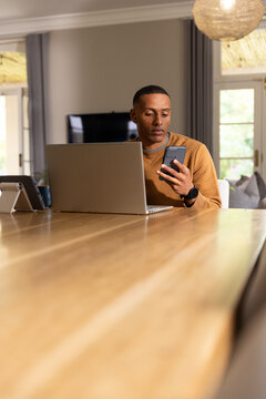 African American man sitting at wooden table wearing rust sweater, holding smartphone, using laptop
