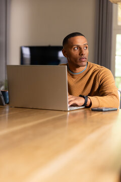 Man in his twenties working on laptop at home dining table with smartphone smartwatch and sweater