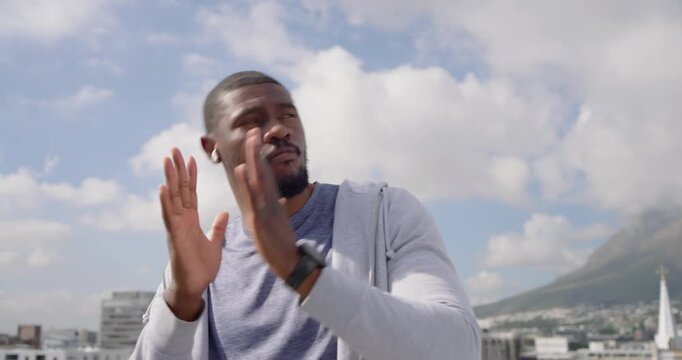 Mid-adult African man wearing earbuds and smartwatch, listening to music, rubbing hands on rooftop
