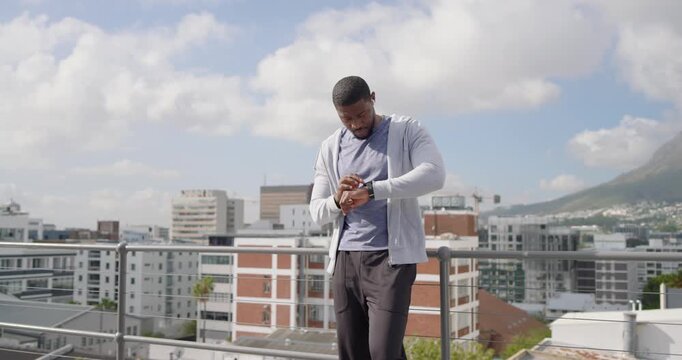 Adjusting earbud, mid adult African man checking smartwatch for alerts, gazing skyline from railing