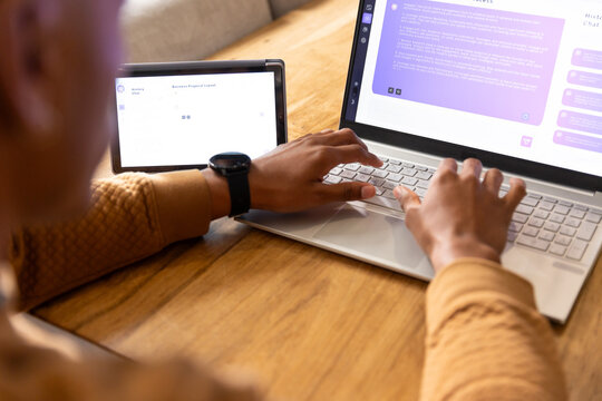 African-American man typing on laptop at wooden table in brown top and smartwatch glancing at phone