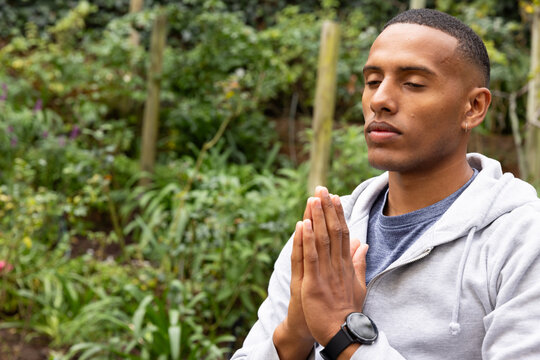 African American man sitting in garden praying, hands pressed, in gray hoodie and watch, copy space